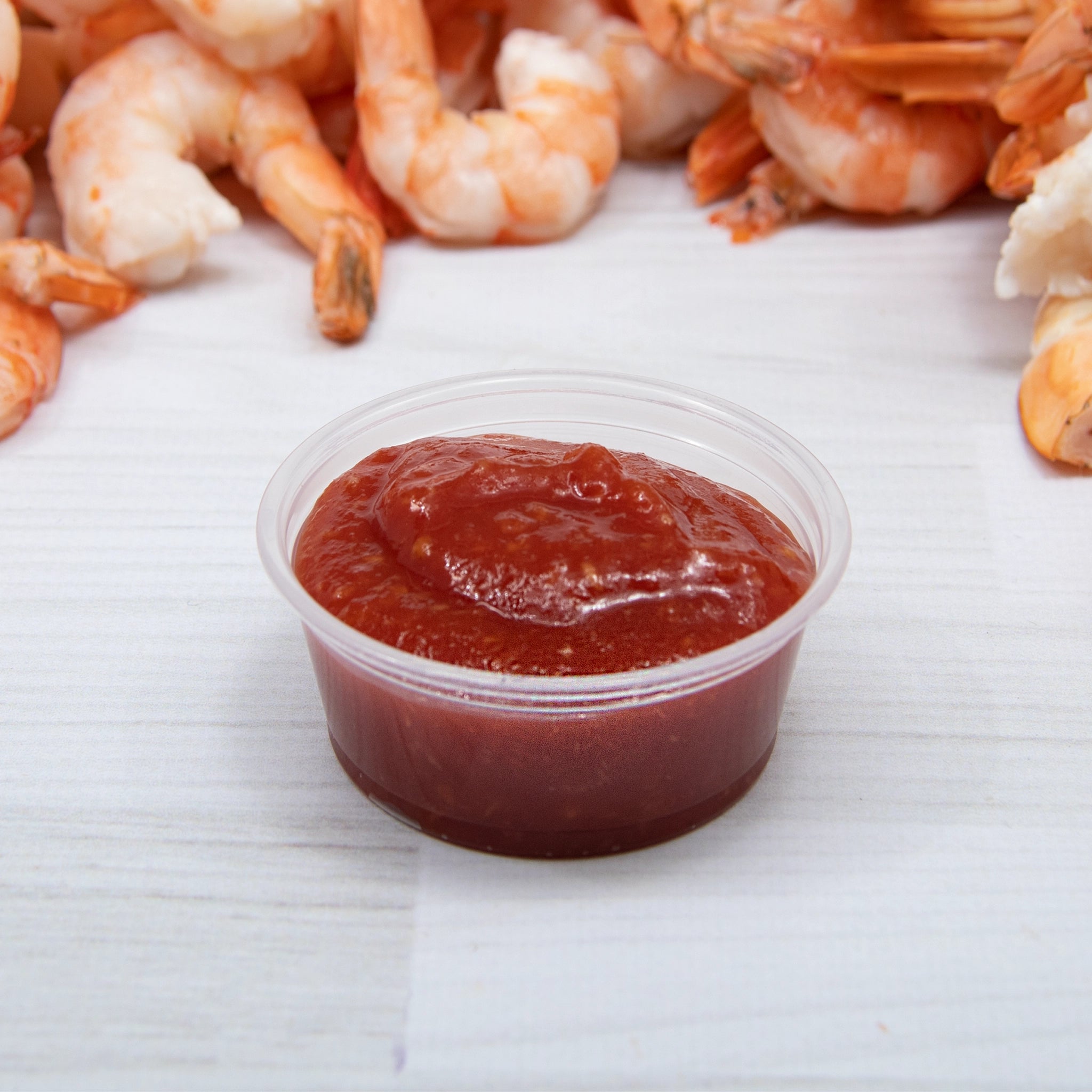 1.5 oz clear plastic portion cup filled with ketchup, surrounded by cooked shrimp on a white wooden surface.