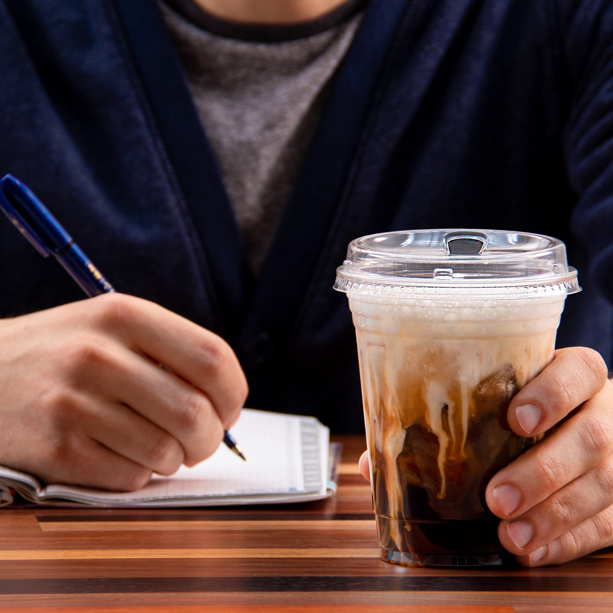 Person holding iced coffee in a 16 oz PET cup sealed with Dart 626NSL sip‑through lid while writing in a notebook.