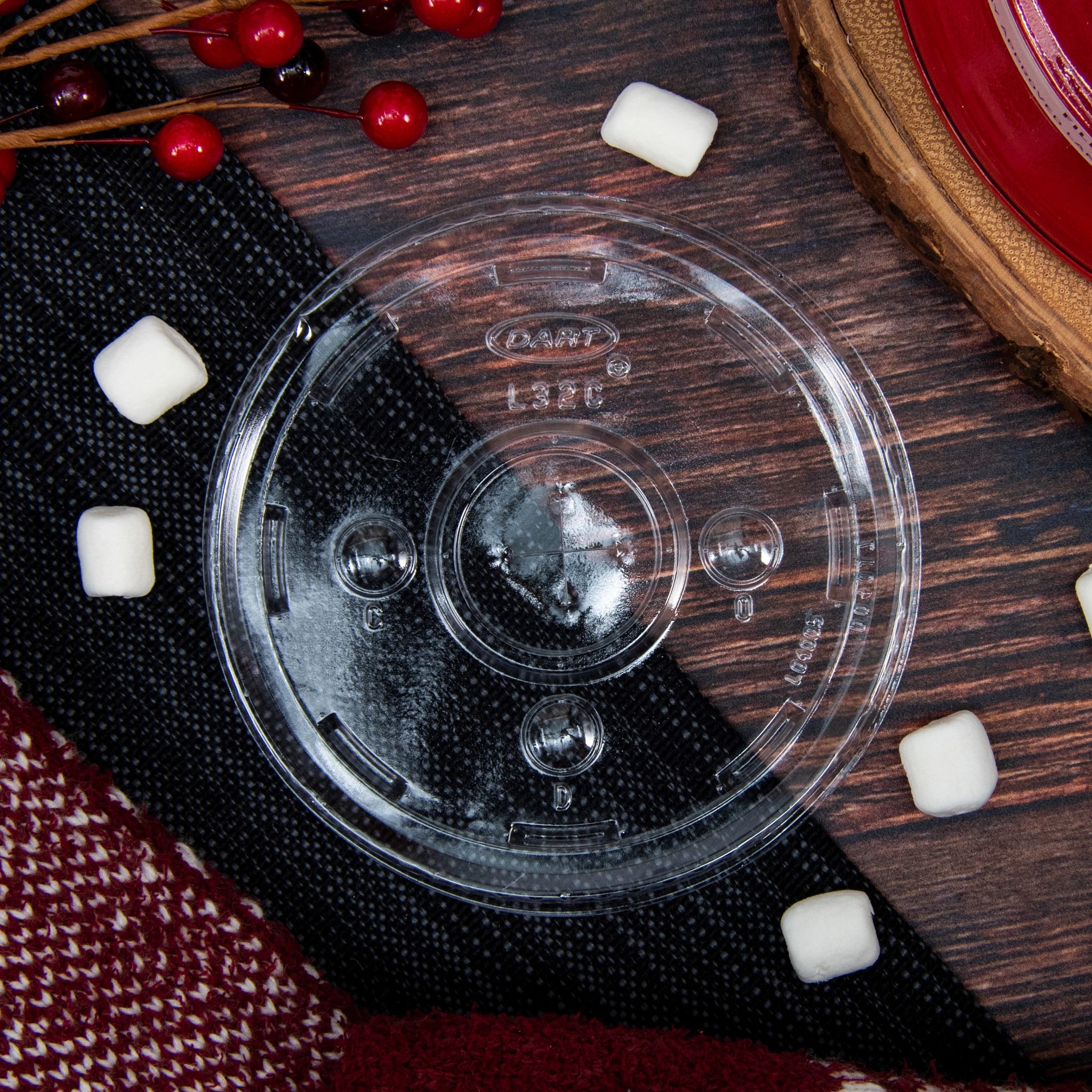 Lifestyle shot of DART L32CR clear flat lid for DART 32AC plastic cups on a wood and fabric surface, surrounded by marshmallows and decorative red berries