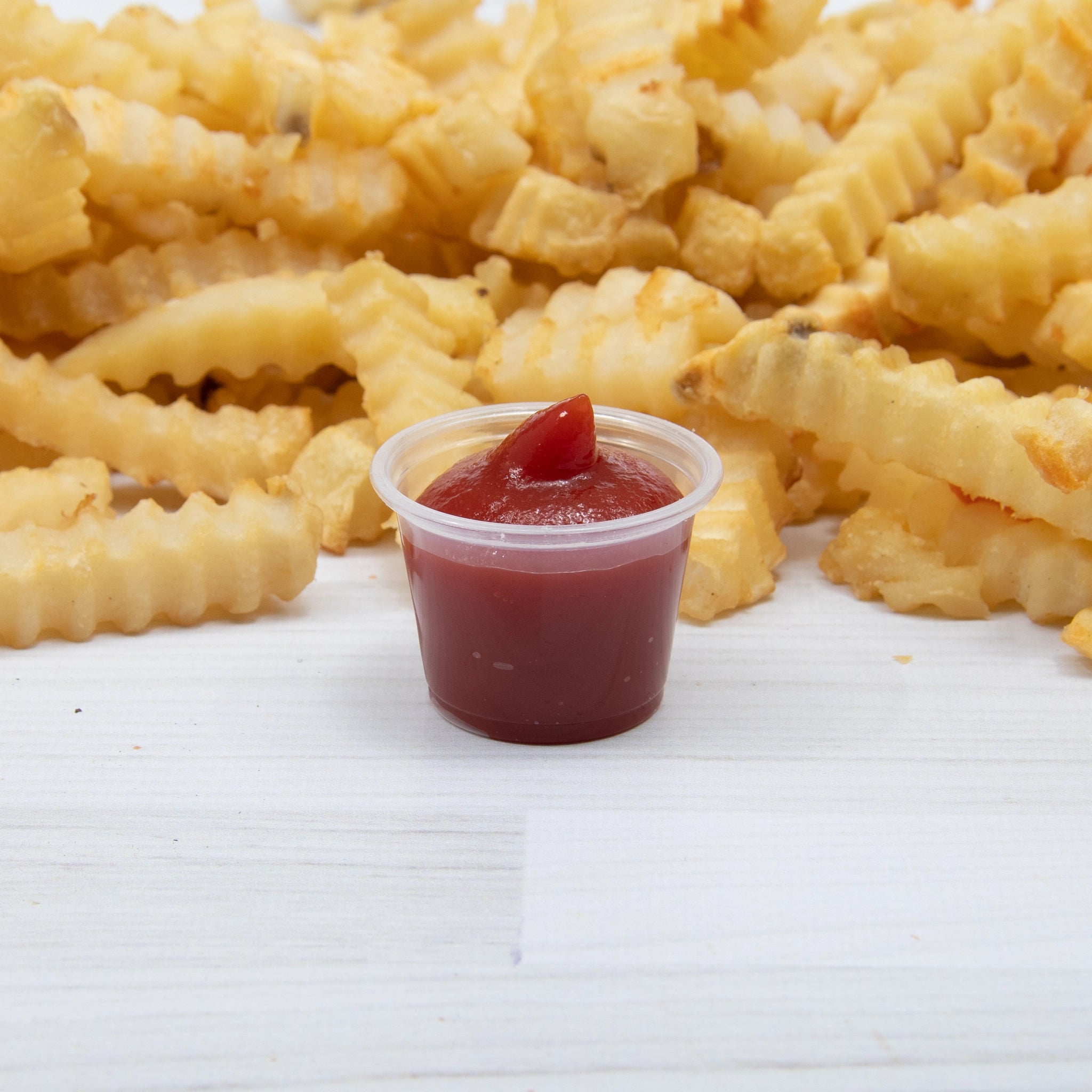 1 oz clear polypropylene plastic portion cup filled with ketchup, surrounded by crinkle-cut fries on a white wooden surface.