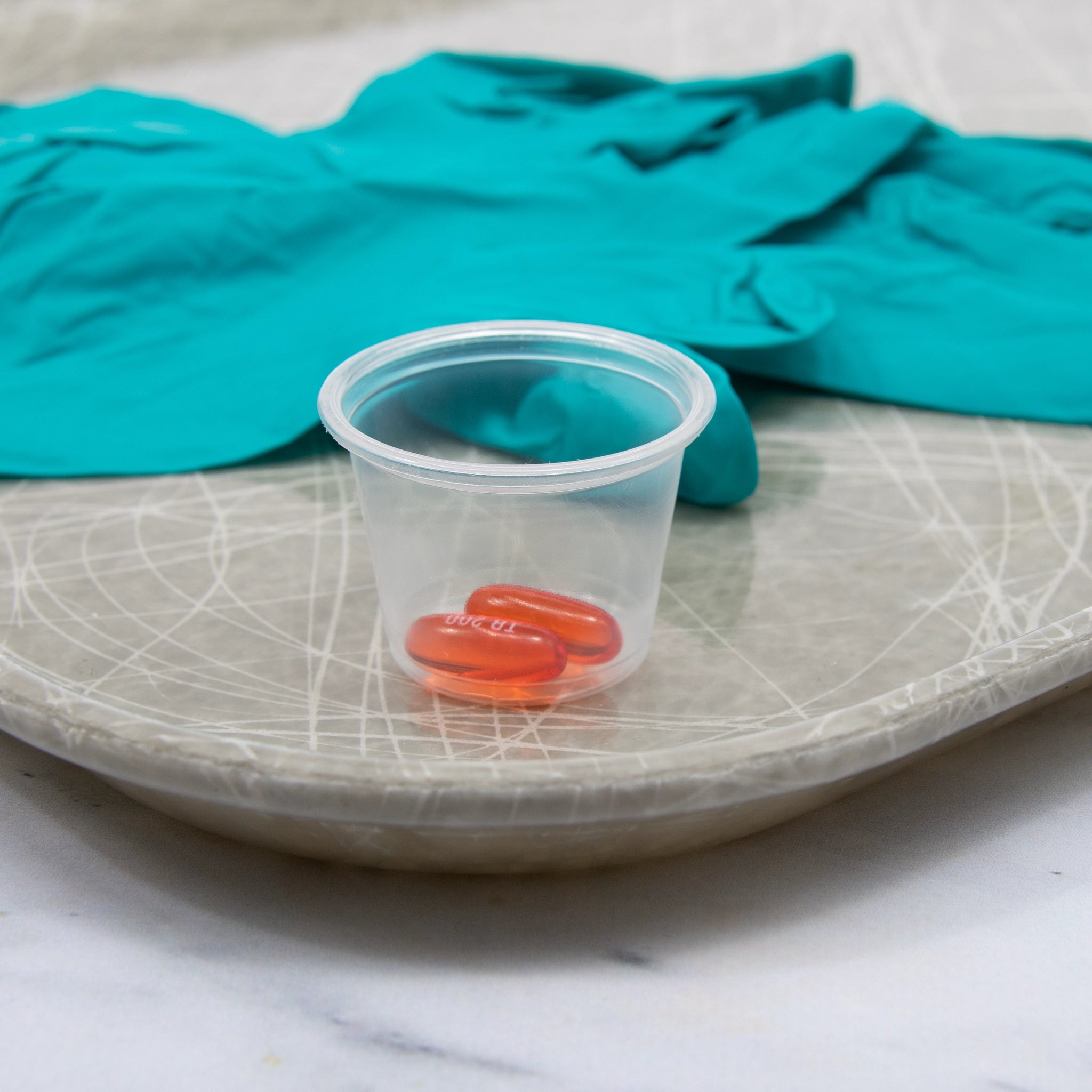 1 oz clear polypropylene plastic portion cup holding red capsule pills, displayed on a gray scratched plate with teal nitrile gloves in the background